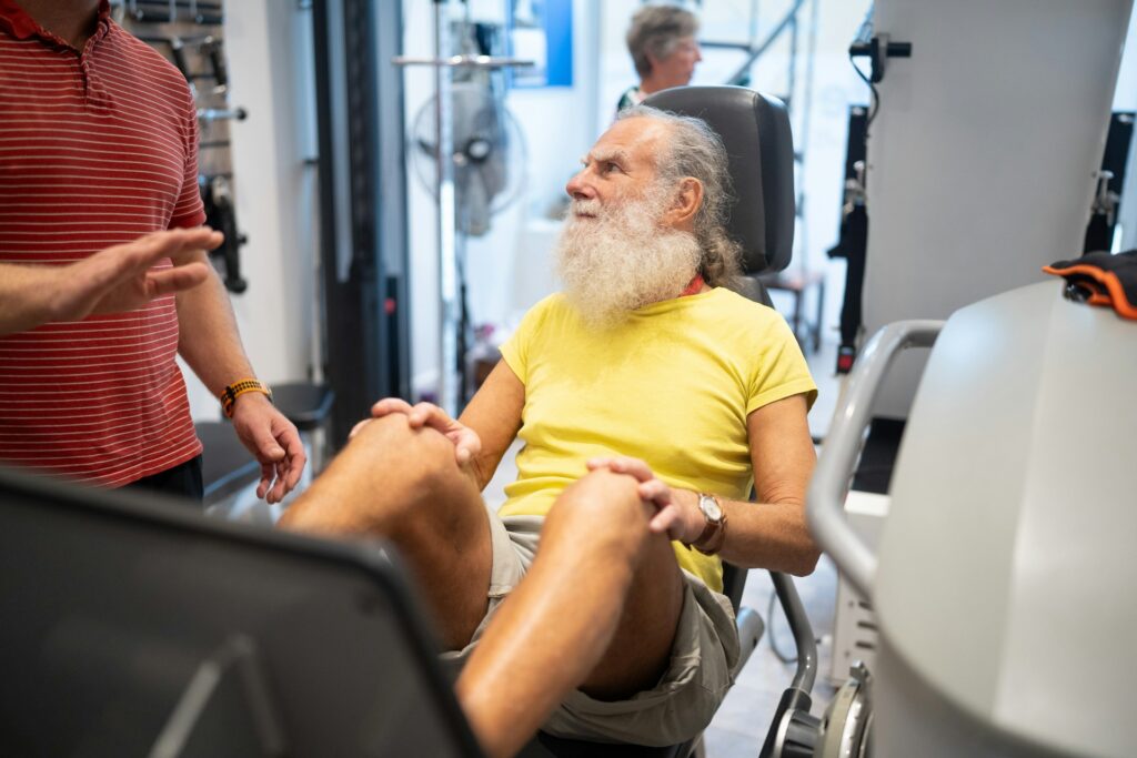Older man with a long white beard doing a leg press machine exercise in a gym, guided by a trainer—emphasizing strength training at any age.
