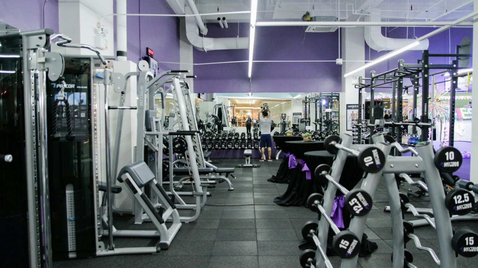 A well-equipped gym with purple walls, featuring various exercise machines, a rack of dumbbells, weight benches, and a person performing an overhead weight exercise in front of a large mirror.